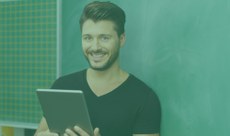 Homem jovem, branco, magro, de cabelo liso e barba sorrindo enquanto segura um tablet prata. Ele usa uma camiseta preta e ao fundo, vê-se uma lousa verde e uma parede ladrilhada.